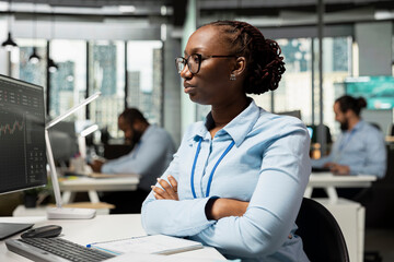 Brokerage company expert in office at workstation desk analyzing stock market charts. African american woman looking at PC financial software interface in prop firm workspace