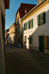 Charming historic street in Dürnstein, Austria, featuring a two-story house with green shutters. Sunlit buildings line the cobblestone street, with a couple walking in the distance under a clear blue 