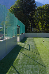 A small soccer ball sits on an artificial turf field enclosed by a green net and fences on a sunny...