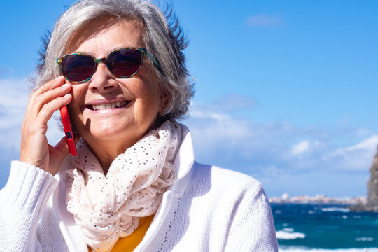 Smiling senior woman talking on a smartphone at the beach. Happy elderly lady wearing sunglasses and scarf enjoying a phone conversation outdoors with the sea and sky in the background. - Powered by Adobe