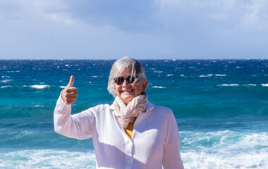 Smiling senior woman wearing sunglasses and scarf enjoying a windy sunny day outdoors at sea...