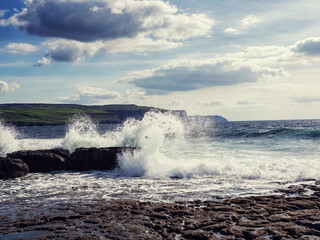 Ragged and tough stone coast hit by power full ocean waves, cliff in the background and cloudy sky. Doolin area, county Clare. Ireland. Popular tourist sightseeing area. Irish nature landscape.