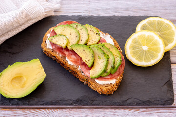 Slice of whole grain bread with seeds with cheese, salami  and avocado for a healthy and tasty snack on a black cutting board. Copy space