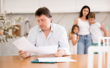 Focused, upset man is sitting at kitchen table and familiarizing himself with studying letter with...