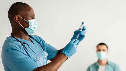 Fighting with outbreak of covid-19, protecting health and immunity against virus. Adult african american doctor in protective mask dials vaccine in syringe for injection to young patient in clinic