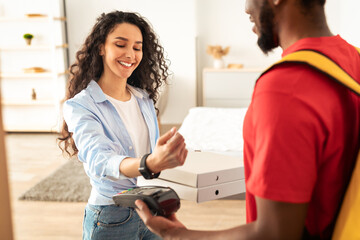 Modern Smart Payment, Food Delivery. Smiling Female Consumer Paying For Pizza With Wristwatch Receiving Boxes From Courier Guy Standing At Door In Her Apartment. Restaurant Delivering Service