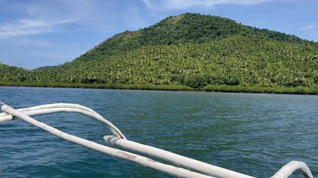 Traditional Philippine banca with outriggers approaches a lush tropical island. Clear turquoise water and palm trees evoke a paradise perfect for Southeast Asian tourism and diving.
