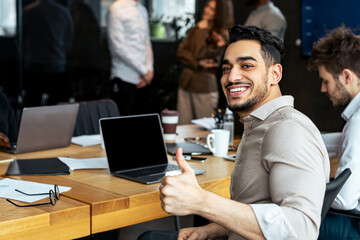 Successful Meeting Concept. Portrait of confident smiling young businessman sitting at desk in modern office with colleagues, showing thumbs up sign gesture, posing and looking at camera