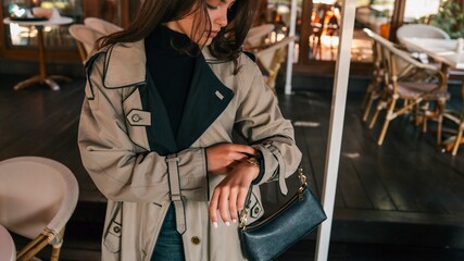 Fototapeta premium Young woman in beige trench coat looking at her wristwatch while standing outside a cozy café, holding a black handbag with a calm, thoughtful expression