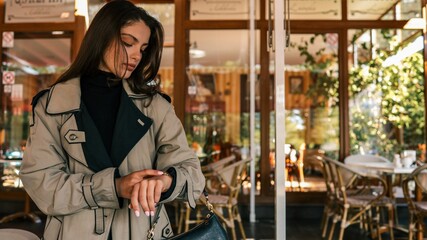 Young woman in beige trench coat looking at her wristwatch while standing outside a cozy café, holding a black handbag with a calm, thoughtful expression