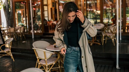 Young woman in trench coat and high-waisted jeans, standing at an outdoor café while adjusting her...