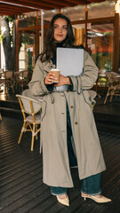Fototapeta premium Elegant woman in trench coat holding a laptop and takeaway coffee cup, standing confidently in a cozy outdoor café setting