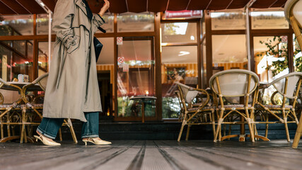Fototapeta premium Low angle view of a fashionable woman walking by a cozy outdoor cafe terrace in a beige trench coat, denim jeans, and heels