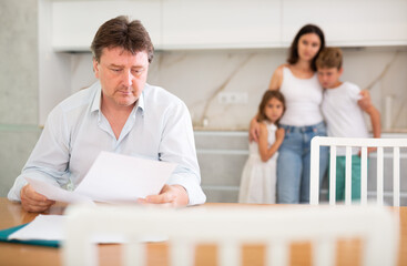 Worried adult man examining documents while sitting at table in kitchen with family