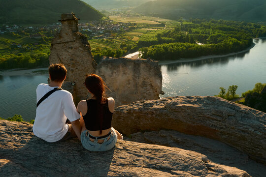 Couple sitting on medieval castle ruins at sunset, overlooking sunlit Wachau Valley, wide Danube River and vineyards, creating romantic and scenic landscape in Austria. - Powered by Adobe