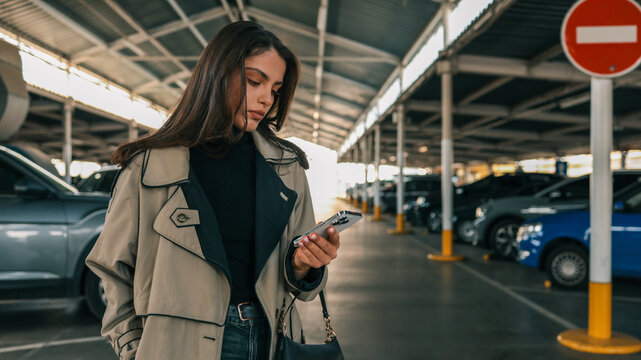 Young woman in beige trench coat using smartphone while standing in covered parking lot with cars