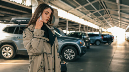 Fototapeta premium Young woman talking on smartphone while standing in a covered parking lot with parked cars. She wears a trench coat and looks serious or focused