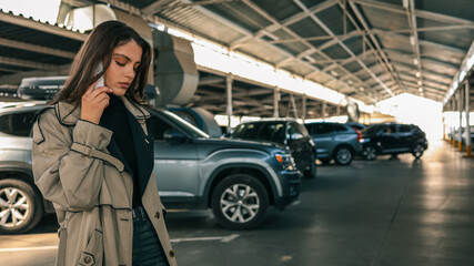 Young woman talking on smartphone while standing in a covered parking lot with parked cars. She wears a trench coat and looks serious or focused © shine.graphics