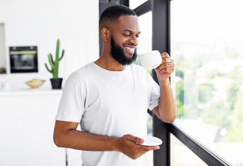 Portrait Of Smiling Relaxed African American Man Enjoying Morning, Drinking Hot Coffee Standing Near Window At Home, Holding Cup And Plate, Starting Day With Favorite Beverage, Copy Space