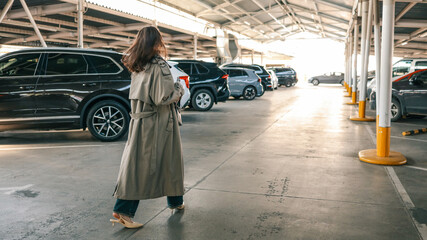 Stylish young woman walking through a covered parking lot, wearing a long trench coat and holding a handbag. She looks over her shoulder while surrounded by parked cars © shine.graphics