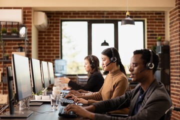 Selective focus on asian woman wearing a headset, seated among diverse coworkers and using computer in brick walled workspace. Multiethnic employees are working at their desks, speaking with clients.