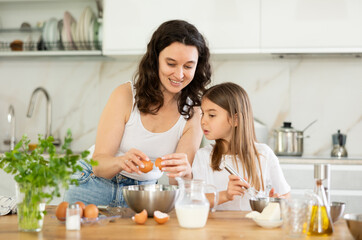 Happy mother and her little daughter are breaking eggs in metal bowl in the kitchen
