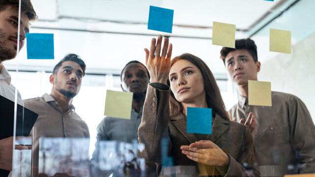 Brainstorm Concept. Creative group of pensive professional diverse people discussing ideas and analysing new business strategy using sticky post-it paper notes. View through transparent glass wall