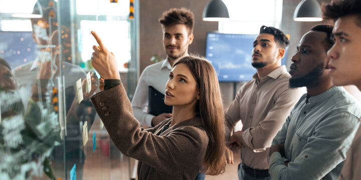 Presentation And Teamwork Concept. Creative group of young professional business people brainstorming, pointing at sticky post-it notes on glass wall in office, discussing new ideas analysing strategy