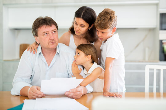 Worried adult man examining documents while sitting at table in kitchen with family - Powered by Adobe