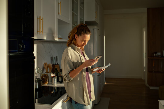 Woman reviewing paperwork and using smartphone in home kitchen