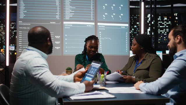 Close up of diverse business staff exchanging reports files and insights to forecast company objectives in a quiet strategy room after hours at night. Finance projections collaboration. Camera A.