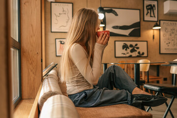 Young woman with long hair sitting on sofa in stylish cafe, holding red cup and enjoying warm drink with modern art posters on wall