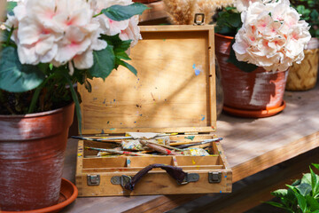Vintage wooden paint box with used brushes and colorful paint stains, placed on wooden table among blooming flowers in pots, illuminated by soft natural sunlight. Art hobby