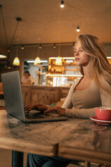 Young woman working on laptop in cozy cafe with warm lighting, cup of coffee on the table, relaxed modern lifestyle scene