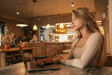 Side view of young woman typing on laptop in cozy cafe interior with warm light and wooden furniture, modern freelance lifestyle scene