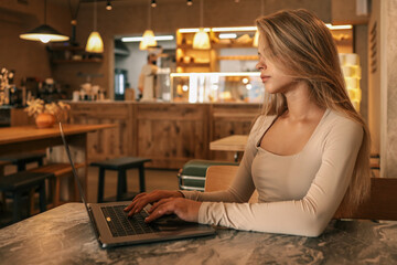 Side view of young woman typing on laptop in cozy cafe interior with warm light and wooden furniture, modern freelance lifestyle scene