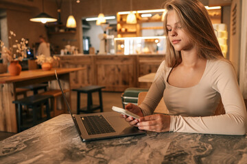 Young woman with long blonde hair using smartphone at cafe table, red coffee cup in foreground, modern cozy interior with warm lights