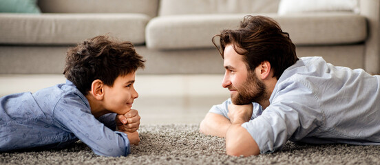 Happy male family. Profile portrait of father and his son lying on carpet, looking at each other and smiling, spending time together at home, side view, panorama © Prostock-studio
