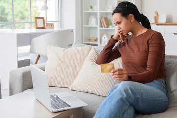 Thoughtful young African-American woman with credit card using laptop on sofa at home