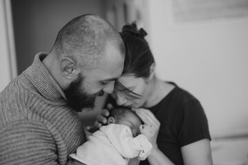 couple with a newborn baby. The father is kissing the mother's forehead while she holds the sleeping baby close. The scene conveys warmth and family love.