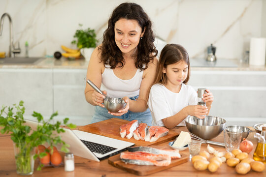 Inexperienced housewife and her daughter participate in online cooking class, cook salmon in batter and watch video instructions on laptop.