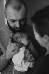 A couple lovingly holds their newborn baby in a cozy indoor setting. The mother gazes at the baby...