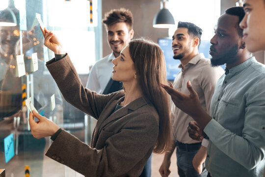 Confident focused female executive writing ideas or tasks on sticky papers on glass wall, young team leader executive manager planning project, organizing work for her male employees