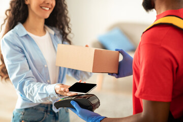 Happy woman using smartphone for contactless payment, black male courier wearing protective gloves...