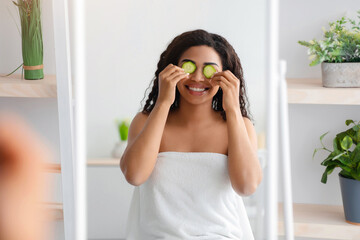 Routine, fun at home, organic vegetables for spa at home. Smiling young african american female in white towel applies pieces of cucumber to eyes, looks in mirror in bedroom interior, empty space