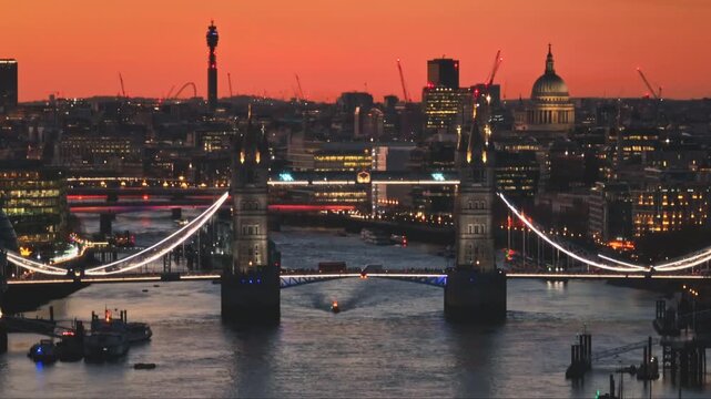 United Kingdom, London: Tower Bridge, illuminated at sunset, majestically spans the River Thames, with St. Paul's Cathedral and the BT Tower enhancing London's skyline. Drone flight footage