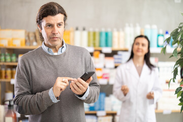 Mature man with a phone in hands flips through an online catalog and looks at the shelves with...