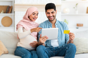 Fototapeta premium Excited pregnant islamic spouses shopping online with tablet and credit card at home while relaxing together on sofa in living room. Happy expectant arab couple buying baby clothes in internet