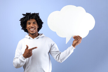 Funky black teen guy pointing at empty speech bubble, smiling at camera on violet studio background, mockup. Handsome Afro teenager with blank word cloud, space for text. News, dialog, announcement