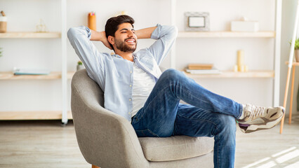 Lazy day. Relaxed arab man resting on comfortable armchair, holding hands behind head. Guy sitting with closed eyes and enjoying weekend in home interior